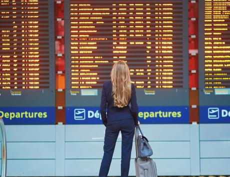 A woman stands in an airport in front of three electronic boards showing current departures. She is wearing a suit and has two bags to symbolise moving to a new location for employment.
